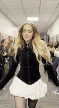 a woman in a black jacket and white skirt is dancing in a hallway with people watching .