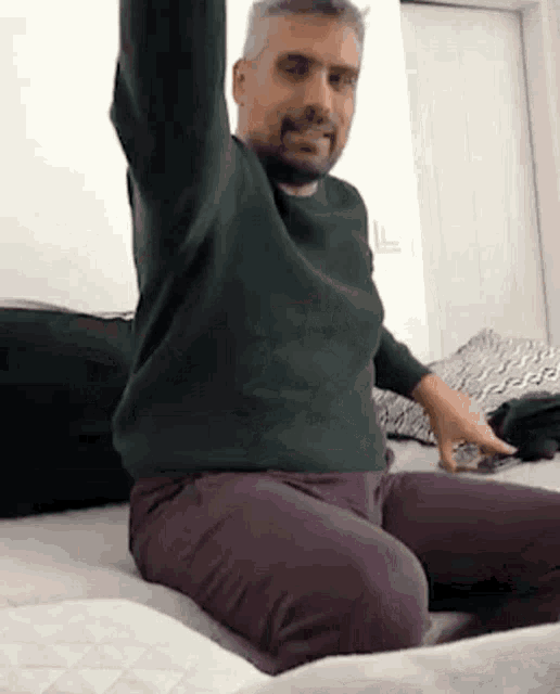 a man in a green sweater and purple pants is sitting on a bed with his arms outstretched .