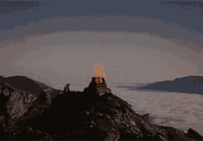 a person standing on top of a rocky mountain with a fire in the background
