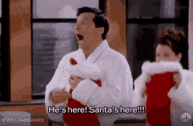a man and a woman are holding christmas stockings and the man is saying he 's here !