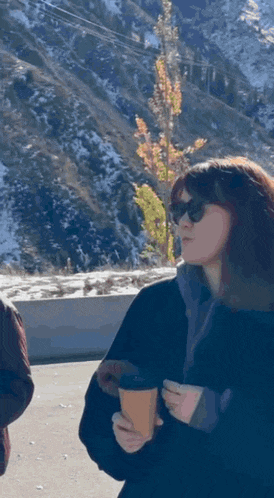 a woman wearing sunglasses is holding a cup of coffee in front of a snowy mountain .