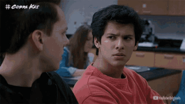two young men are sitting at a table in a classroom talking to each other .