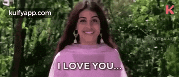 a woman is smiling and saying `` i love you '' while standing in front of a forest .