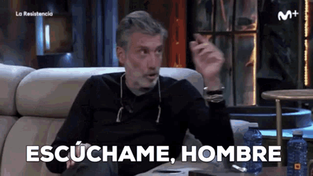a man with a beard is sitting on a couch and saying " escuchame hombre "