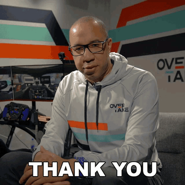a man wearing a hoodie that says overtake is sitting on a couch and says thank you