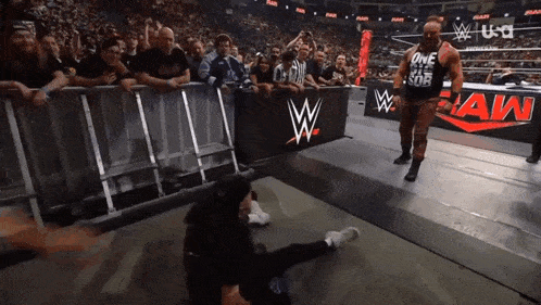 a man in a black shirt with the word one on it stands in front of a wrestling ring