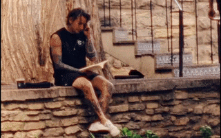 a man with tattoos is sitting on a stone wall reading a book