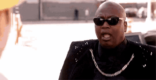 a bald man wearing sunglasses and a chain is talking to a man .