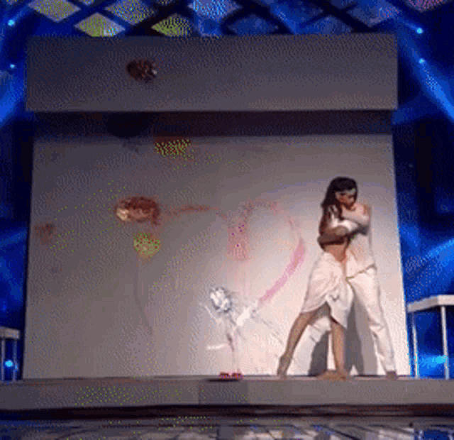 a man and a woman are dancing on a stage in front of a wall with a heart drawn on it