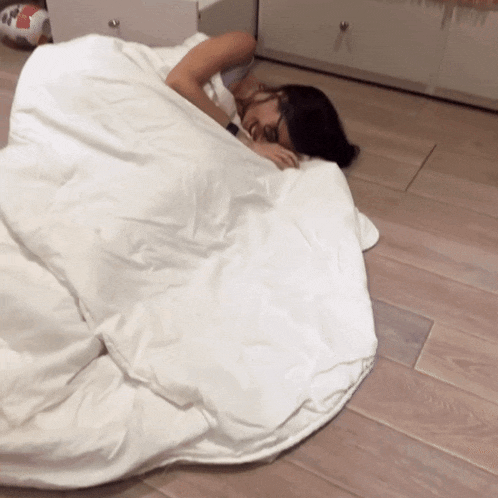 a woman laying under a white blanket on a wooden floor