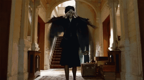 a woman in a black dress with wings is standing in a hallway covering her face .