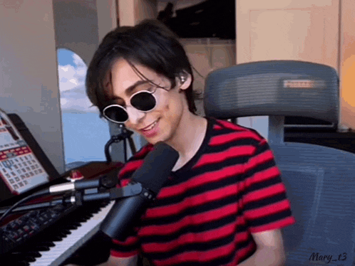 a young man wearing sunglasses and a striped shirt is sitting at a piano and singing into a microphone .