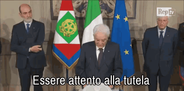 a man in a suit and tie stands in front of flags and says " essere attento alla tutela " at the bottom