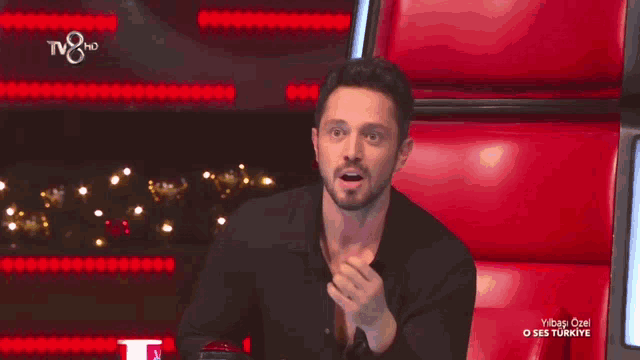 a man in a black shirt is sitting on a red chair with tv8 hd in the background