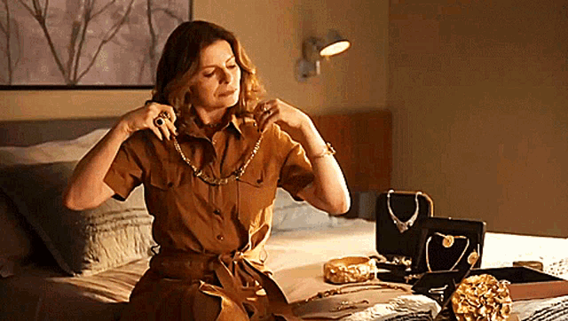 a woman in a brown dress is sitting on a bed looking at her jewelry
