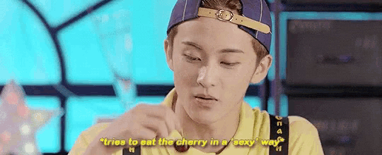 a young man wearing a hat and a yellow shirt is eating a cherry in a sexy way .
