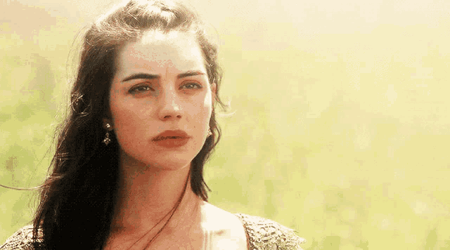a woman with dark hair and earrings stands in a field of grass