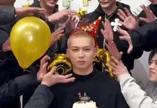a man wearing a party hat is surrounded by people holding balloons .