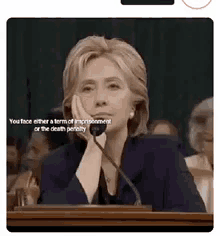 hillary clinton is sitting at a podium with her hand on her chin