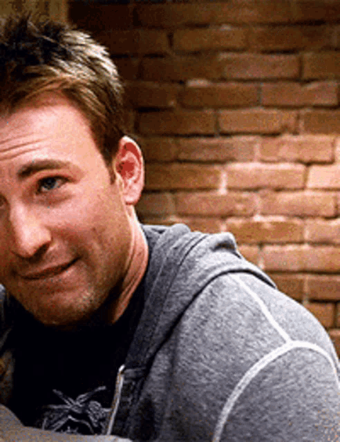 a man wearing a grey hoodie is standing in front of a brick wall and smiling