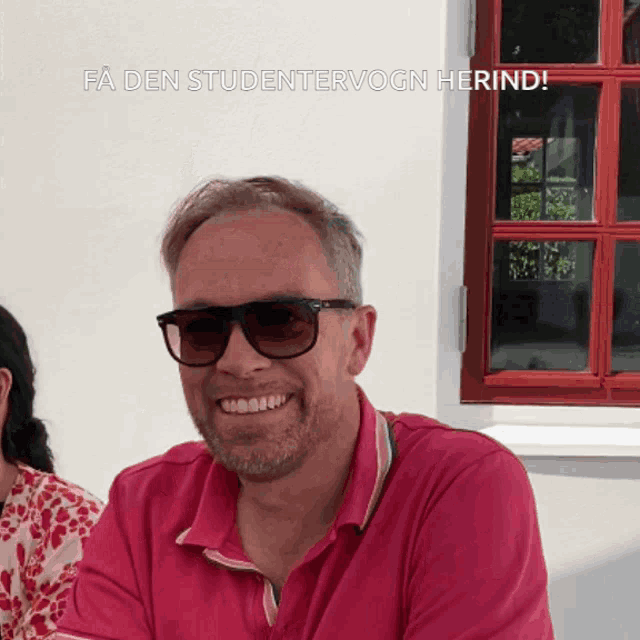 a man wearing sunglasses and a pink shirt smiles in front of a window with the words fa den studentervogn herind below him