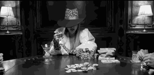 a woman in a cowboy hat is sitting at a table with poker chips and drinking a martini .