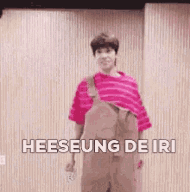 a man in overalls is standing in front of a wooden wall and says heeseung de iri .