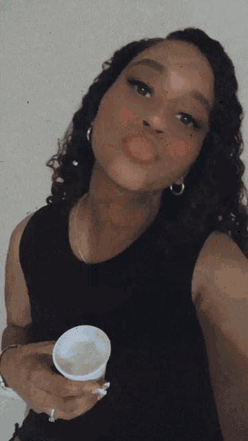 a woman in a black tank top is holding a white container and sticking her tongue out
