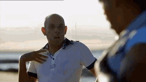 a bald man in a white polo shirt is standing on a beach talking to another man .