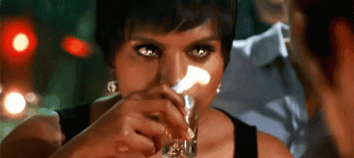 a woman is drinking a shot of whiskey from a glass while a man looks on .