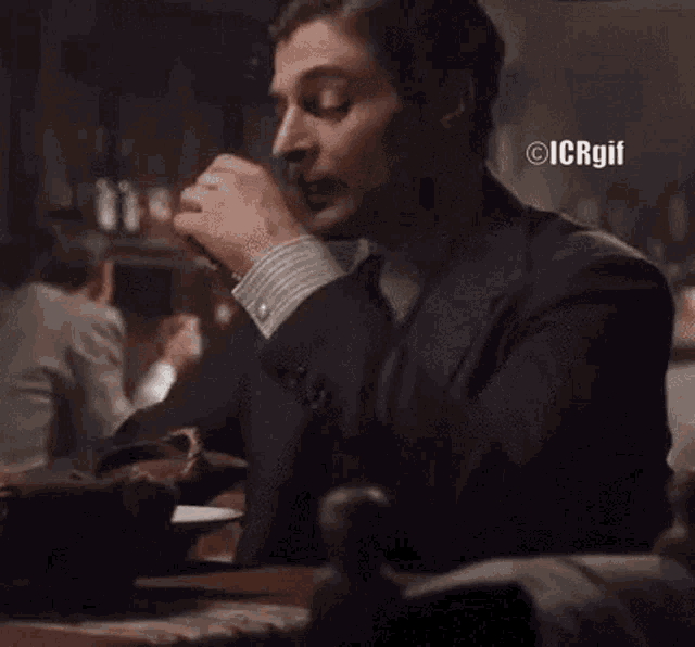 a man in a suit and tie is sitting at a table smoking a cigarette in a dark room .