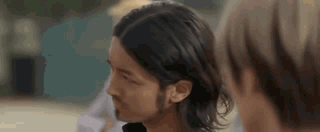 a man with long hair and a beard is talking to another man in a blurry photo .