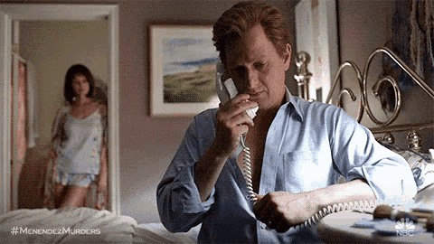 a man is talking on a telephone in a bedroom while a woman stands in the background .