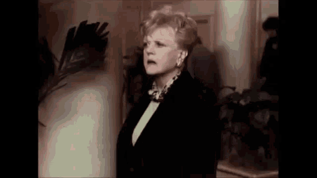 a woman in a black suit and a necklace is standing in a hallway .