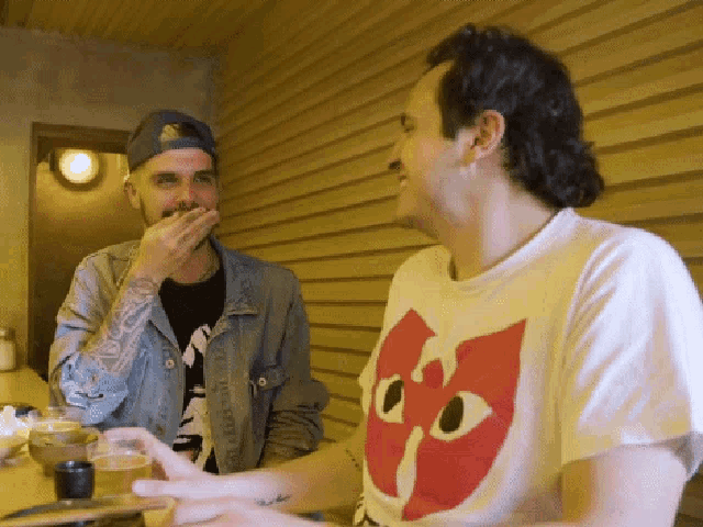 two men are sitting at a table with one wearing a shirt that has a cat on it