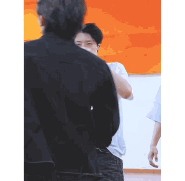 a man in a black jacket is standing next to another man in a white shirt in front of an orange wall .