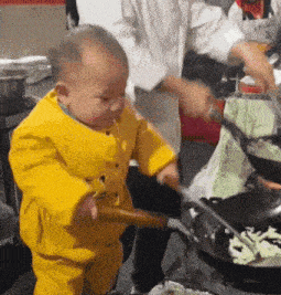 a baby in a yellow outfit stirs food in a wok