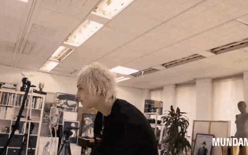 a man with blonde hair is standing in a room with a skeleton in the background and the word mundan in the corner .
