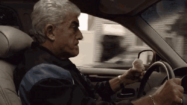 a man in a blue and black jacket is driving a car with his hand on the steering wheel