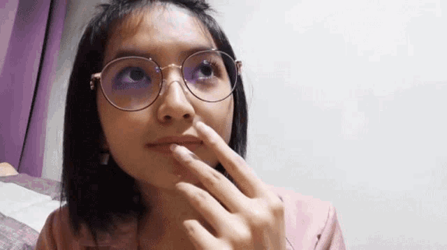 a woman wearing glasses and a pink shirt is touching her lips with her finger .