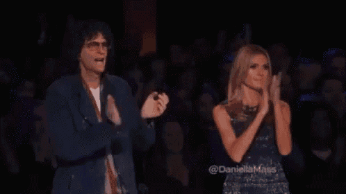 a man and a woman are clapping in front of a crowd . the woman is wearing a sequined dress .