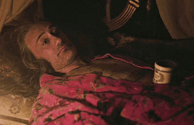 a man with a beard is laying under a pink blanket next to a mug that says " coffee "