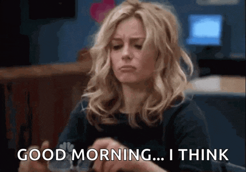 a woman is sitting at a table and saying `` good morning ... i think '' while holding a cup of coffee .