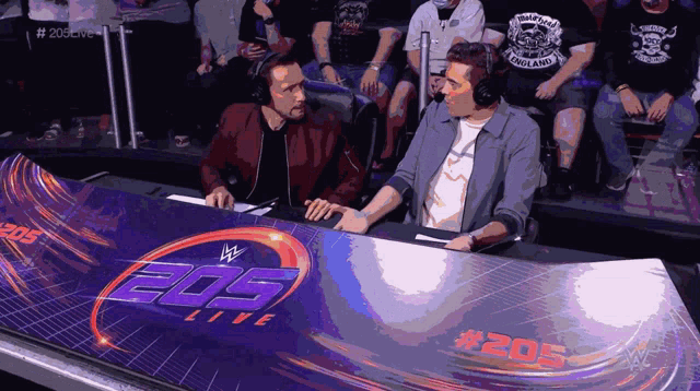 two men are sitting at a table that says 205 live on it