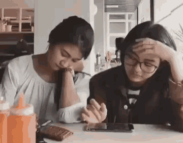 two women are sitting at a table with their hands on their foreheads looking at a cell phone .