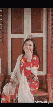 a woman in a red dress is sitting on a wooden chair covering her mouth with her hand .