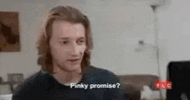 a man with long hair is sitting in a living room talking to someone and says `` pinky promise ? ''