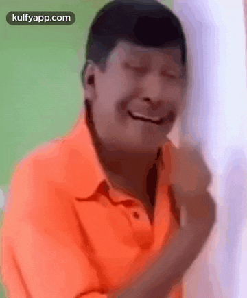 a man in an orange shirt is laughing with his eyes closed and his hands in his pockets .