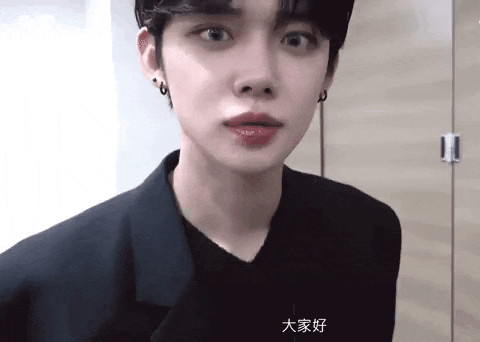a young man wearing a black jacket and earrings is standing in front of a wooden door .