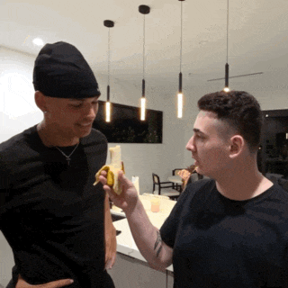 a man in a black hat is eating a banana from another man 's hand